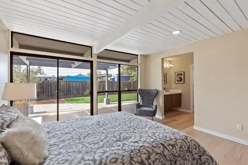 Bright bedroom with patterned bedding, armchair, and large sliding glass doors opening to a backyard patio.
