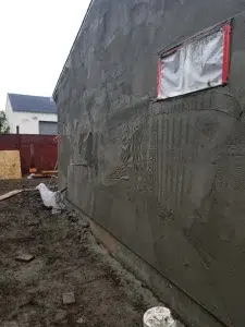 Fresh stucco applied to an exterior wall with a masked window and wet ground along the building’s foundation.