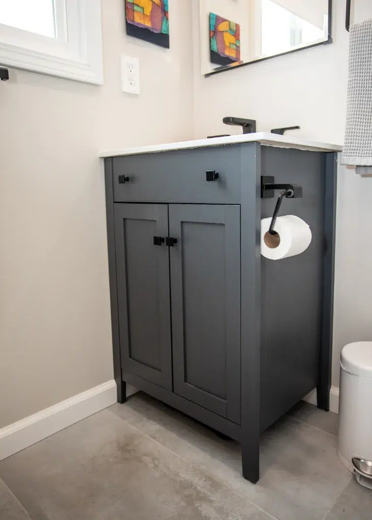 Gray bathroom vanity with white countertop, black faucet, and wall-mounted toilet paper holder.