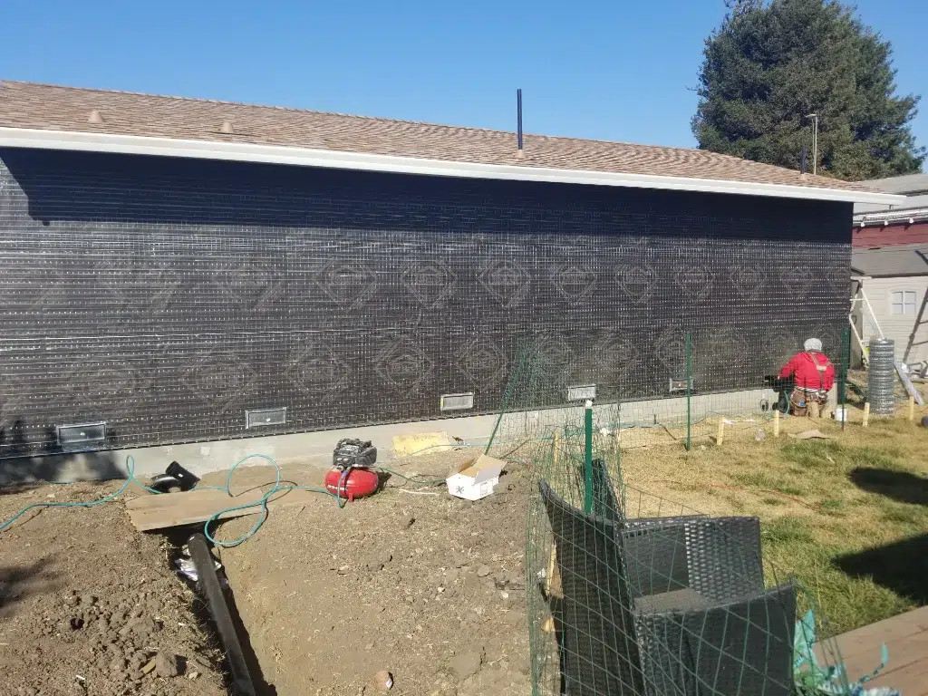 Worker installing metal lath over black stucco paper on a long exterior wall with tools and fencing materials nearby.