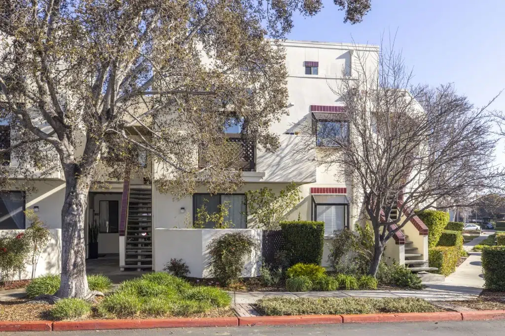 Modern beige apartment building with red brick accents, exterior stairs, and landscaped trees along the sidewalk.