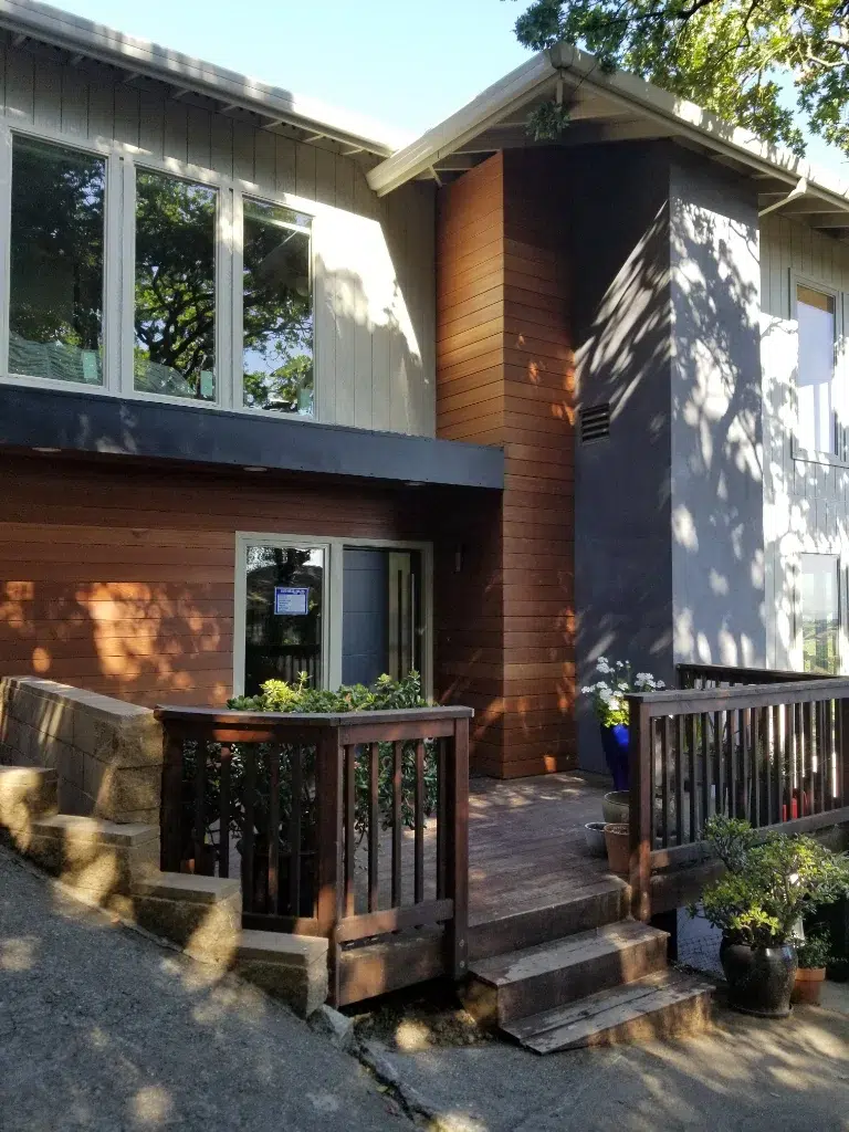 Two-story home with wood and gray siding, large front windows, and a small wooden entry deck with potted plants.