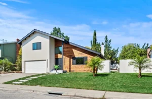 Modern suburban house with wood and stucco exterior, green lawn, and palm trees under a clear blue sky.
