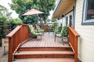 Raised wooden deck with steps, outdoor chairs with green leaf cushions, and a red patio umbrella next to a house