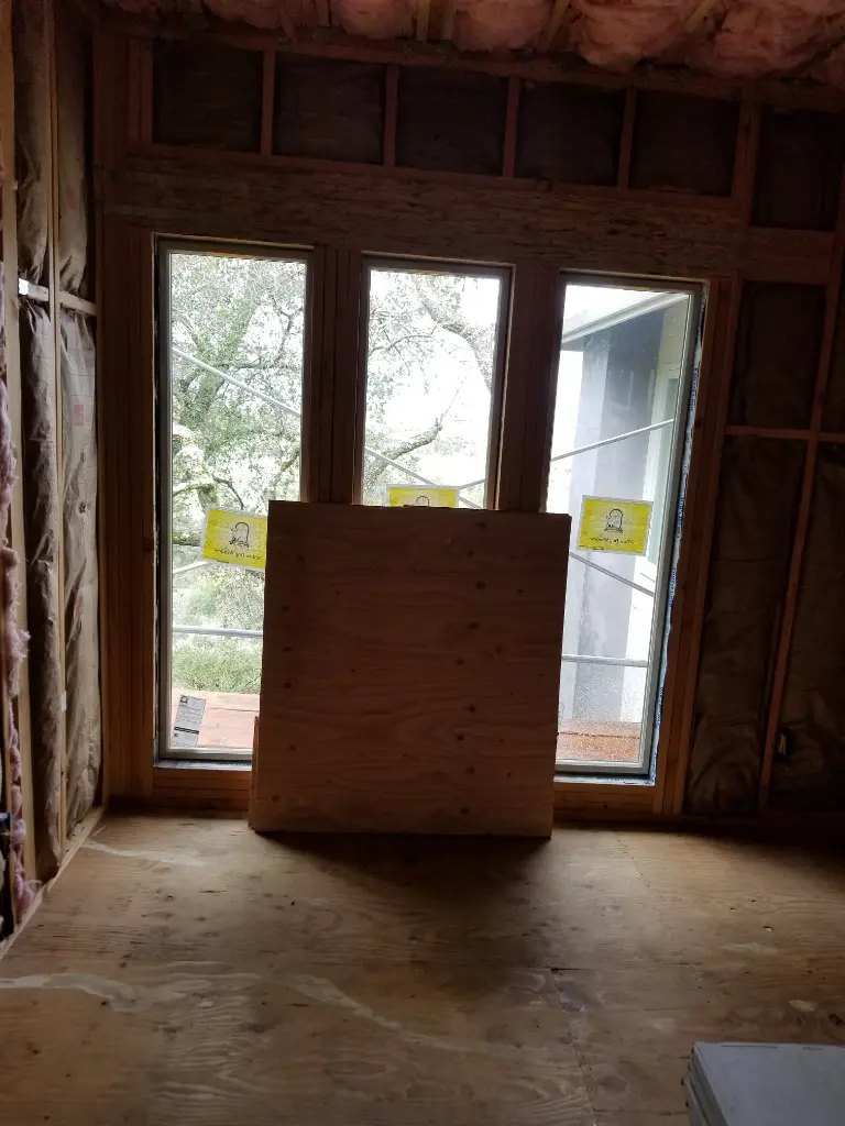 Unfinished interior room with exposed insulation, three vertical windows, and a plywood board covering the lower center window