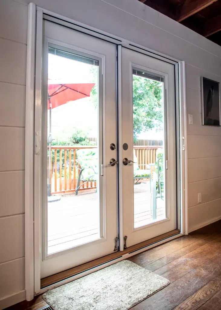 White French doors opening to a wooden deck with patio furniture and a red umbrella.